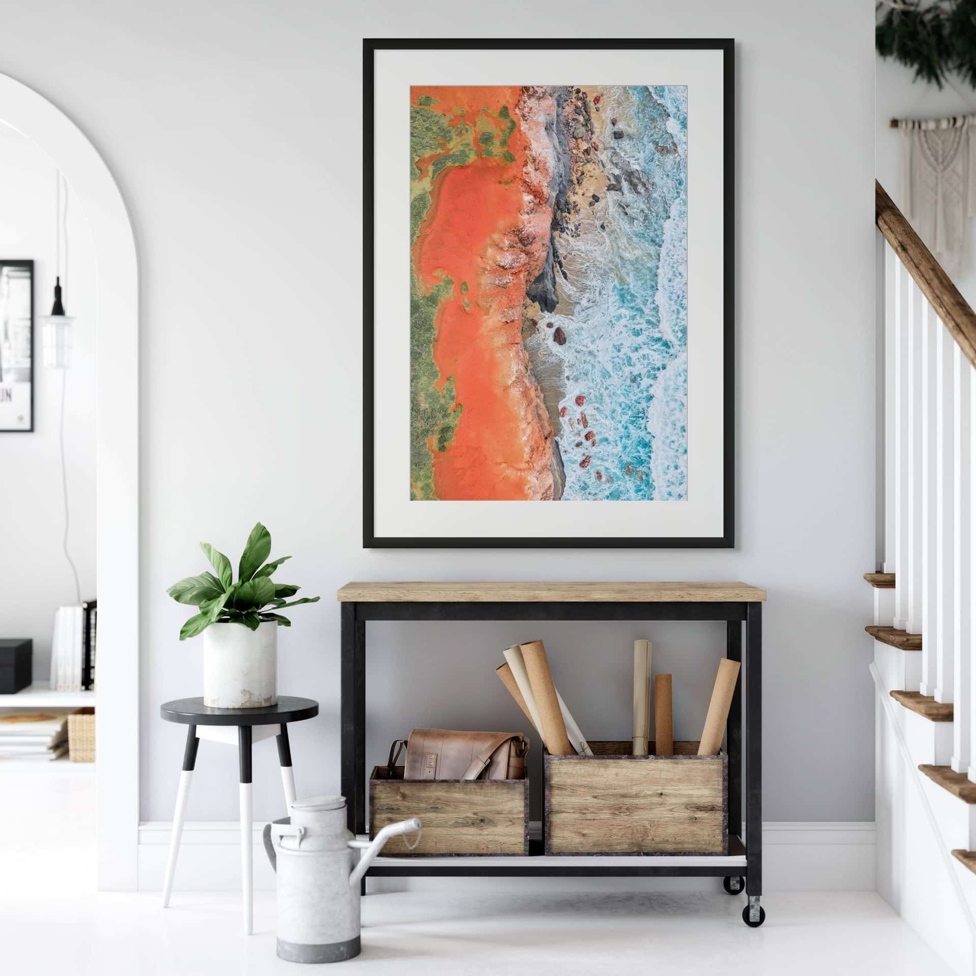 A modern entrance with a console table, baskets, and rolled posters, plus a plant on a round side table. Above hangs THE SEAM, a limited edition fine art piece featuring Australian photography in red, brown, and blue tones. - The Coastal Edit Australia