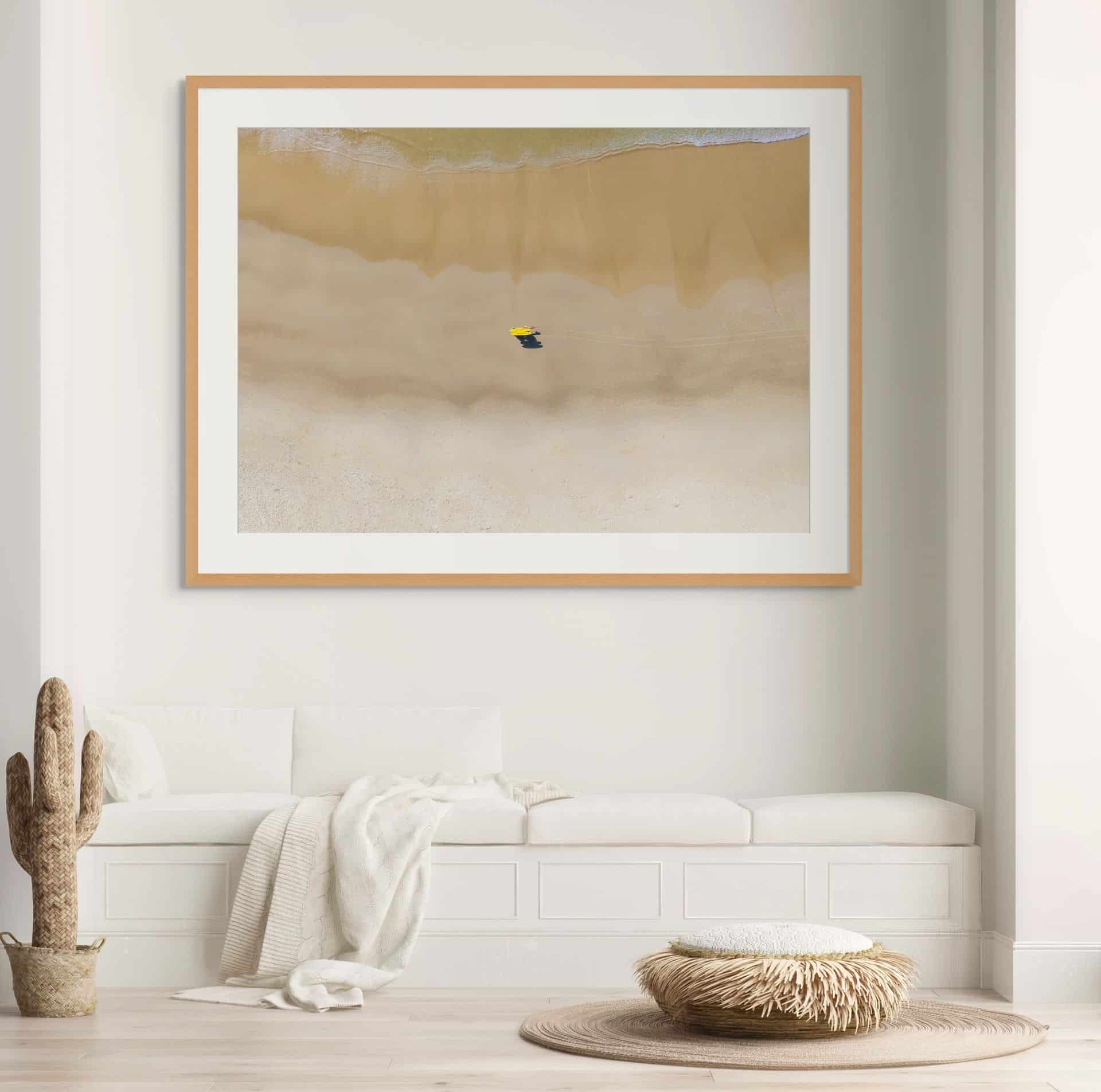 The framed limited edition fine art aerial photo "VIGILANCE" shows a sandy beach, gentle waves, and a person under a yellow umbrella, displayed above a white bench in a minimalist, neutral-toned lounge with cactus and woven decor. - The Coastal Edit Australia