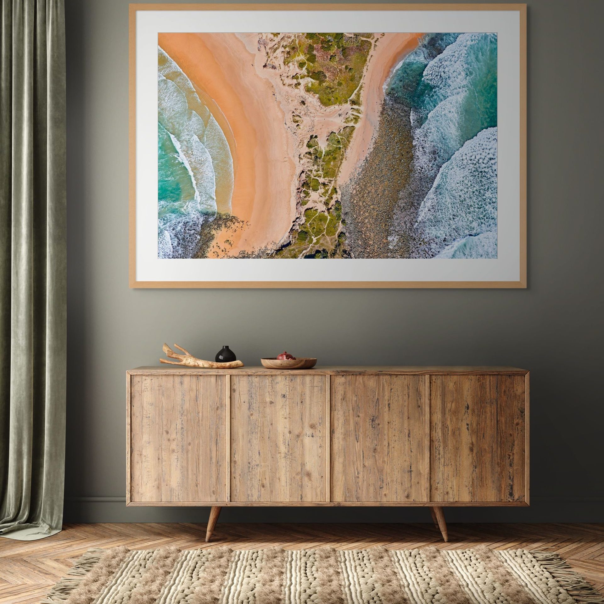A large framed aerial photo titled "BIFURCATE," a limited edition Australian fine art piece, hangs above a wooden sideboard with décor on a geometric rug. A grey wall and partially visible curtain complete the setting. - The Coastal Edit Australia