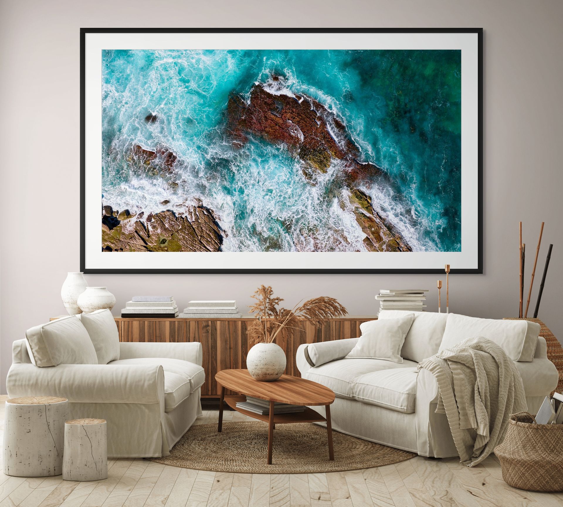 A modern living room with white sofas, a wooden coffee table, and neutral decor. Above the sofas hangs THE CONSTANT: a limited edition aerial photograph of waves over rocks in museum-grade framing. - The Coastal Edit Australia