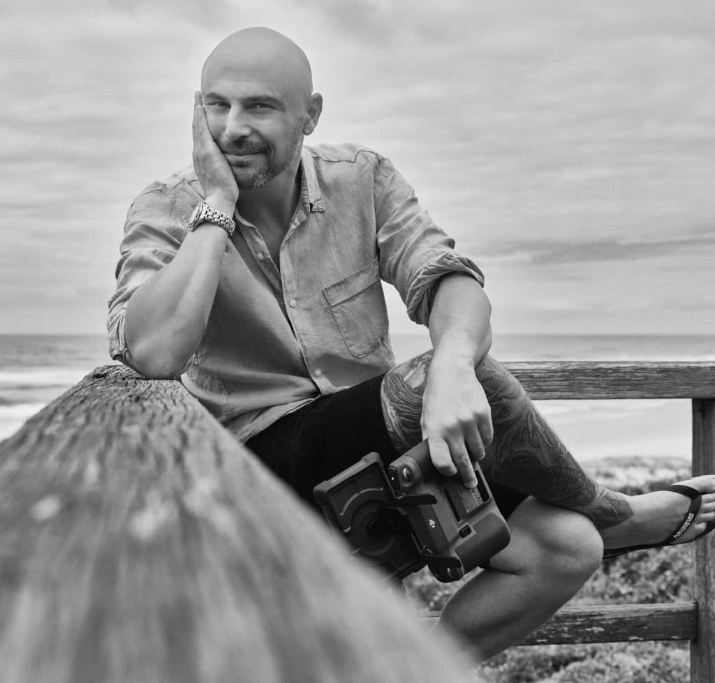 A bald man with a beard sits on a wooden rail by the beach, resting his head on his hand and holding a camera. He wears a casual shirt, shorts, and has a tattoo on his leg. This black and white shot evokes Australian photography. - The Coastal Edit Australia