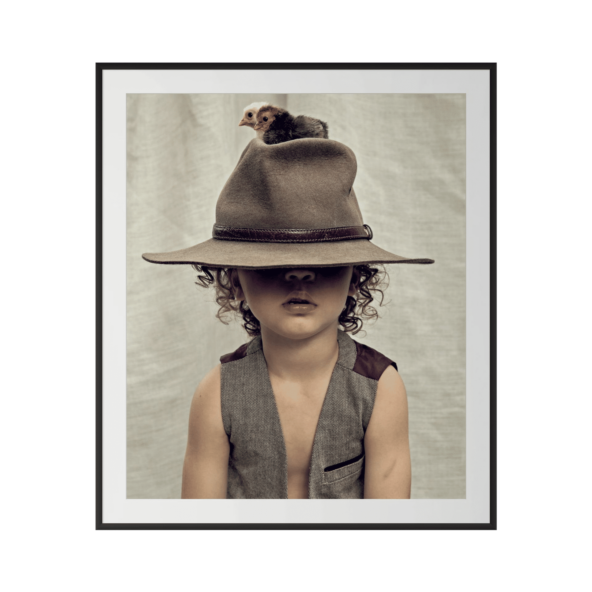 A young child in a sleeveless vest and wide-brimmed hat—eyes concealed—sits beneath a chick on the hat, framed by a neutral backdrop. BLOODLINE is ideal wall art to bring charm to luxury coastal interiors. - The Coastal Edit Australia
