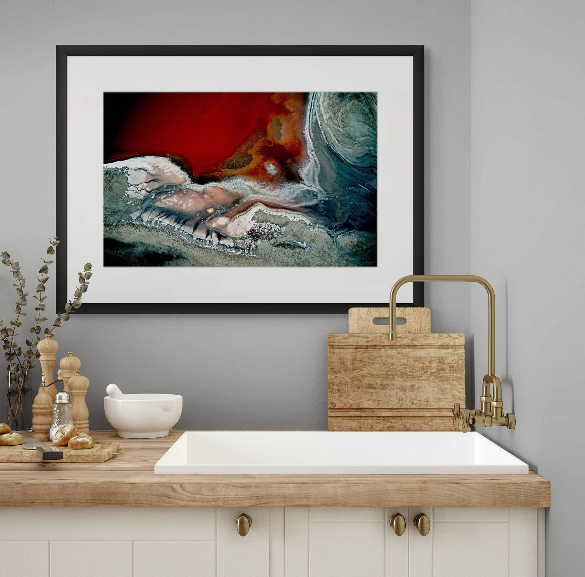 A modern kitchen with light wood worktops, a white sink, brass tap, and chopping boards features CRIMSON TIDE, a limited edition fine art piece in red, white, and blue earth tones above the sink. Dried plants complete the look. - The Coastal Edit Australia