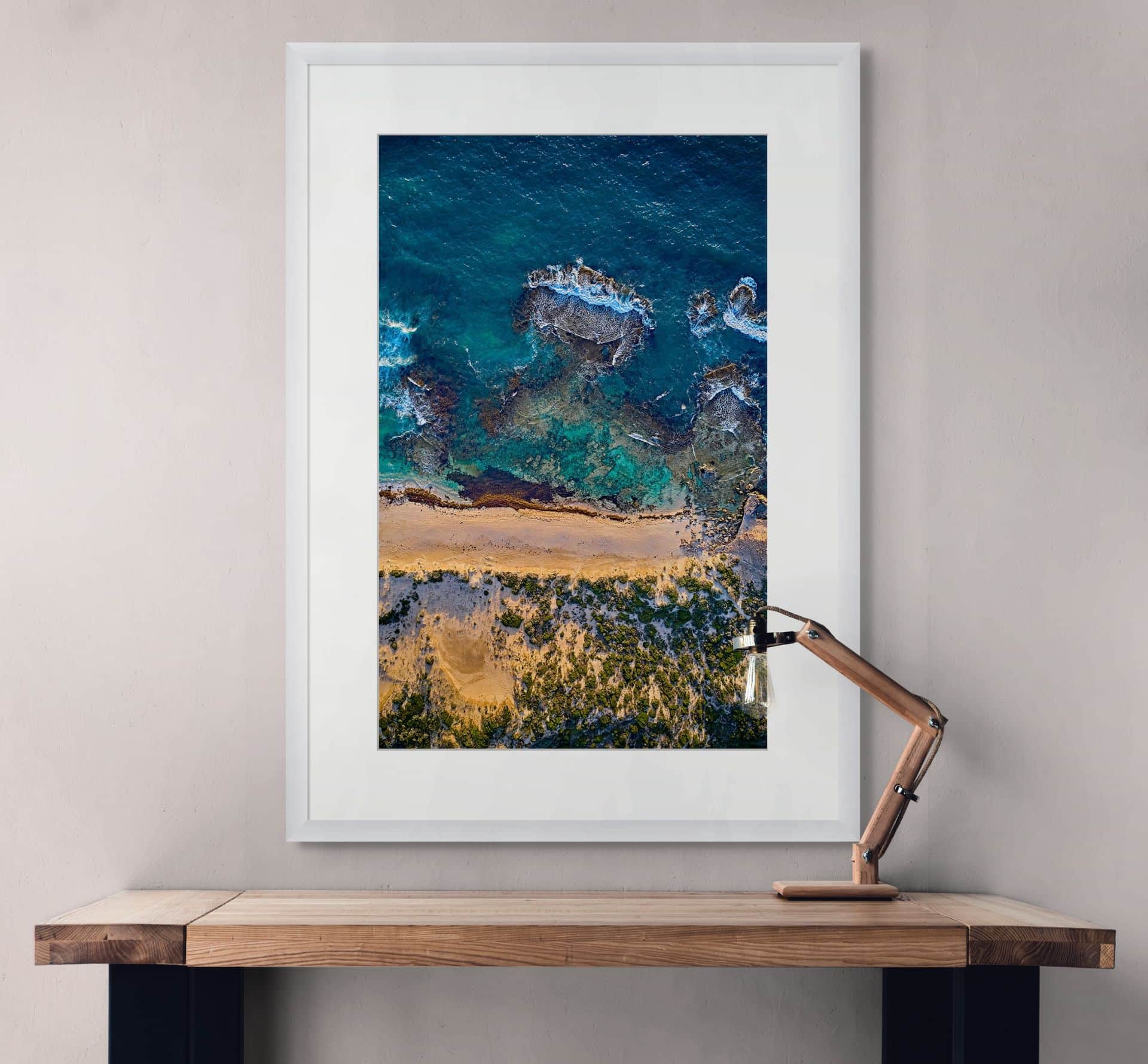 AEOLIAN, a framed aerial photo of a coastline with sandy beach, rocks, and blue-green sea, hangs above a wooden desk—a striking piece of Australian photography ideal for interior design wall art. - The Coastal Edit Australia
