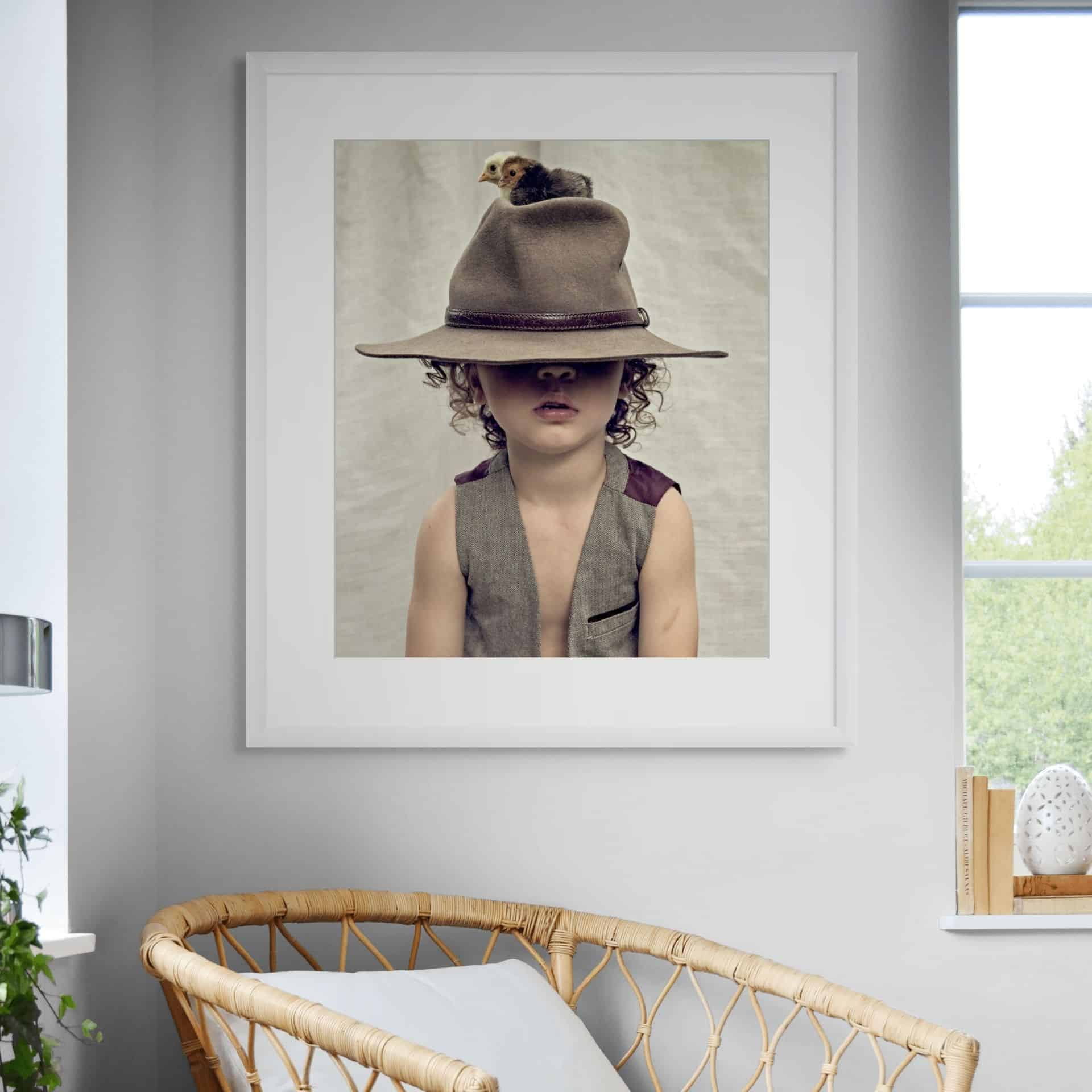 BLOODLINE: A museum-grade framed photo of a child in a large hat with a chick on top, wearing a sleeveless waistcoat above a wicker chair in a bright room, exemplifying Australian photography. - The Coastal Edit Australia